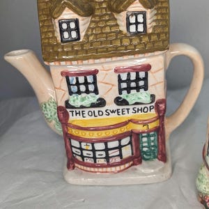 May include: A ceramic teapot shaped like a small house with a brown roof and a sign that reads "The Old Sweet Shop". The teapot is decorated with windows, a door, and a candy shop interior.