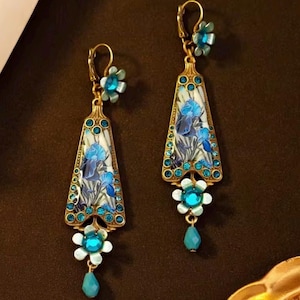 Peut inclure: Une paire de boucles d'oreilles dorées avec un motif floral. Chaque boucle d'oreille présente un long pendentif triangulaire avec une peinture d'iris bleu, entouré de cristaux de couleur turquoise. Un charme de fleur et de goutte pend du bas.