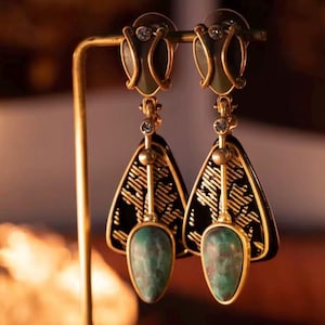 Könnte beinhalten: Ein Paar goldfarbene Ohrringe mit einem schwarz-goldenen Filigranmuster. Jeder Ohrring hat einen tropfenförmigen, grünen Edelstein. Die Ohrringe werden auf einem goldfarbenen Ohrringständer präsentiert.