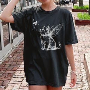 May include: A black short-sleeve t-shirt featuring a white graphic print of a kitten with fairy wings looking up at a small butterfly surrounded by tiny stars. The design is centered on the chest area of the garment, which has a crew neckline.