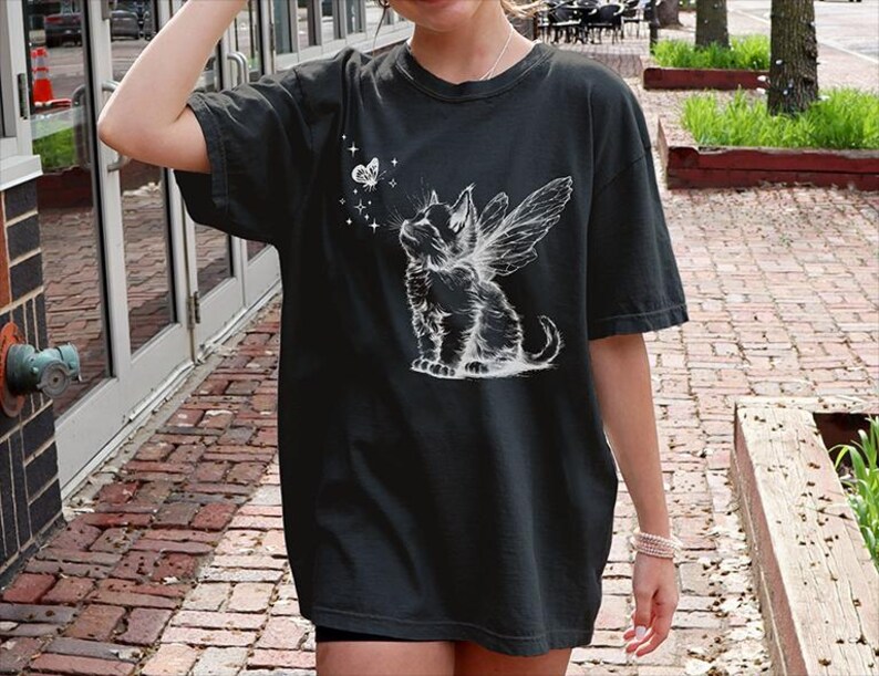 May include: A black short-sleeve t-shirt featuring a white graphic print of a kitten with fairy wings looking up at a small butterfly surrounded by tiny stars. The design is centered on the chest area of the garment, which has a crew neckline.