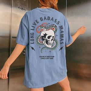 May include: A blue t-shirt with a graphic of a skull with a snake wrapped around it. The text "Long Live Badass Mamas" is printed on the back of the shirt. The text "Doin' it better since day one" is printed below the graphic.