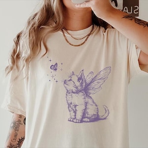 May include: A white t-shirt with a purple illustration of a cat with wings and a butterfly. The cat is looking up at the butterfly.