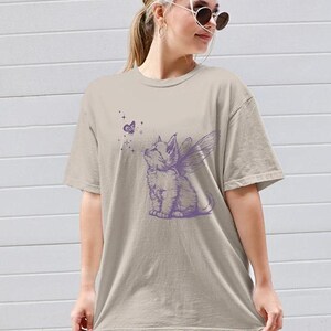 May include: A beige short-sleeved t-shirt featuring a purple graphic print of a kitten with fairy wings looking up at a small butterfly surrounded by tiny stars. The shirt has a crew neckline and a relaxed, loose-fitting silhouette.