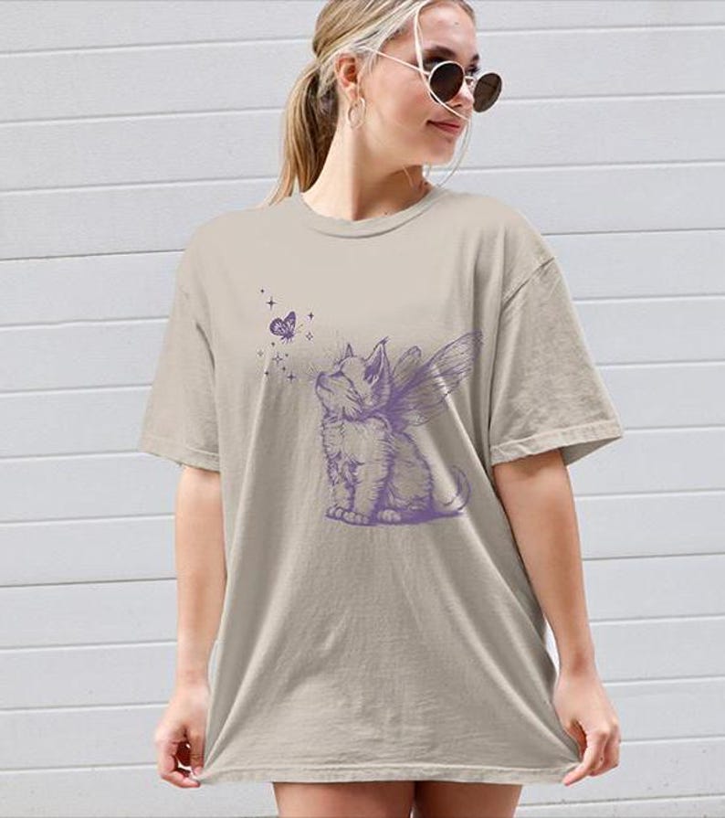 May include: A beige short-sleeved t-shirt featuring a purple graphic print of a kitten with fairy wings looking up at a small butterfly surrounded by tiny stars. The shirt has a crew neckline and a relaxed, loose-fitting silhouette.