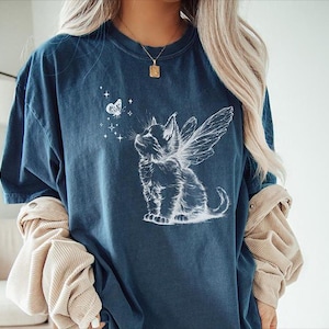 May include: A dark blue t-shirt featuring a white line art graphic of a kitten with fairy wings looking up at a small butterfly surrounded by tiny stars. The shirt has a crew neckline and short sleeves, worn over a beige corduroy long-sleeve shirt.