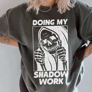May include: A black t-shirt with a white graphic of a skeleton with a hood, giving a thumbs up. The text "Doing My Shadow Work" is printed below the graphic.