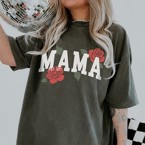 May include: Dark olive green t-shirt with the word "MAMA" in white block letters, flanked by red rose illustrations with green leaves. The shirt has a relaxed fit and short sleeves, perfect for casual wear.