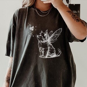 May include: A charcoal grey short-sleeve t-shirt featuring a white graphic print of a kitten with fairy wings looking up at a small butterfly surrounded by tiny stars. The shirt has a relaxed fit and a crew neckline, showcasing a detailed illustration.