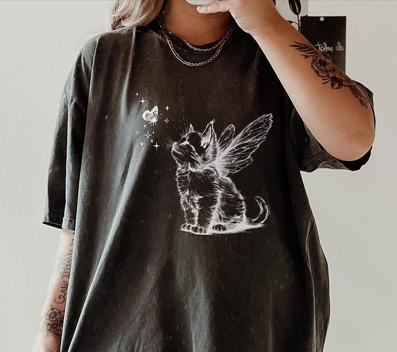 May include: A charcoal grey short-sleeve t-shirt featuring a white graphic print of a kitten with fairy wings looking up at a small butterfly surrounded by tiny stars. The shirt has a relaxed fit and a crew neckline, showcasing a detailed illustration.