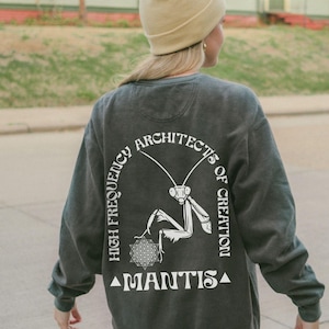 May include: A charcoal gray long-sleeve sweatshirt featuring a white graphic on the back. The design displays a praying mantis, a geometric flower of life pattern, and the text "HIGH FREQUENCY ARCHITECTS OF CREATION" and "MANTIS" in a bold, stylized font.