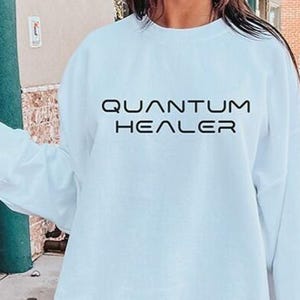 May include: A light blue crewneck sweatshirt featuring the text "QUANTUM HEALER" printed in a bold, black, modern sans-serif font across the chest. The garment has long sleeves, a ribbed neckline, and a relaxed fit, providing a simple design.