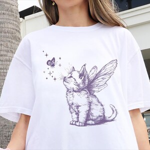 May include: A white t-shirt featuring a purple graphic print of a kitten with fairy wings looking up at a small butterfly surrounded by four-pointed stars. The illustration is rendered in a detailed sketch style with fine lines and shading.