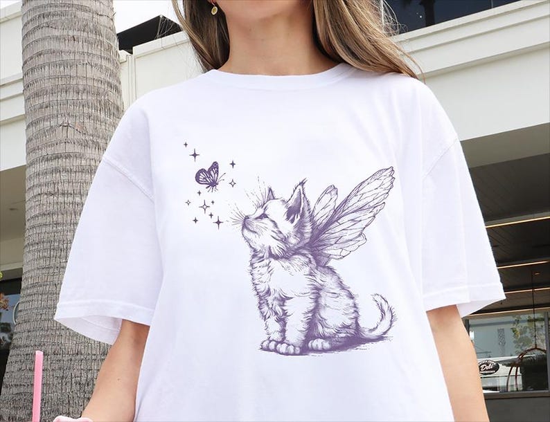 May include: A white t-shirt featuring a purple graphic print of a kitten with fairy wings looking up at a small butterfly surrounded by four-pointed stars. The illustration is rendered in a detailed sketch style with fine lines and shading.