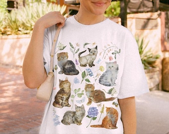 May include: A white short-sleeve t-shirt featuring a colorful watercolor-style graphic print of seven kittens in various poses, interspersed with delicate botanical illustrations of green leaves, small blue flowers, and purple blossoms.