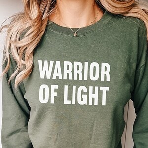 Peut inclure: Un sweat-shirt vert à manches longues avec le texte "WARRIOR OF LIGHT" imprimé en blanc sur le devant.