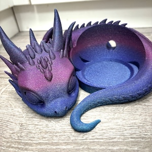 Könnte beinhalten: Eine schlafende Drachenfigur mit einem Farbverlauf von Lila und Blau. Der Drache hat eine strukturierte Oberfläche und ein kleines Loch im Rücken.