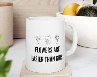 Może przedstawiać: Biały ceramiczny kubek z czarnym napisem "FLOWERS ARE EASIER THAN KIDS" i trzema małymi ilustracjami kwiatów. Kubek stoi na beżowej podstawce.