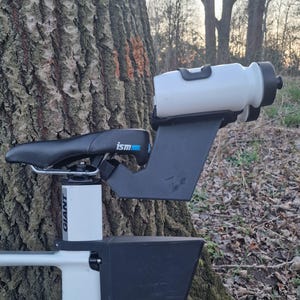 Puede incluir: Una bicicleta blanca y negra con un asiento negro y un portabotellas. La botella de agua es blanca con una tapa negra. La bicicleta está apoyada en un árbol.