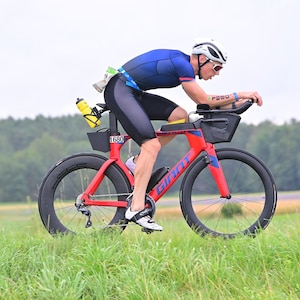 Könnte beinhalten: Eine Person in einem blauen und schwarzen Radtrikot fährt ein rotes Triathlon-Rad der Marke Giant mit schwarzen Rädern und einem schwarzen Lenker. Das Fahrrad hat einen gelben Wasserflaschenhalter und eine schwarze Aufbewahrungstasche am Rahmen. Die Person trägt einen weißen Helm und eine Sonnenbrille.