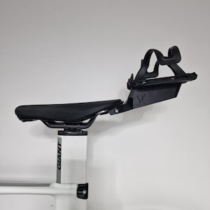 May include: A black bicycle saddle mounted on a white Giant seat post, featuring an attached black rear bottle cage mount with a stylized logo. The assembly includes a matte black water bottle cage secured to the rear bracket behind the seat.