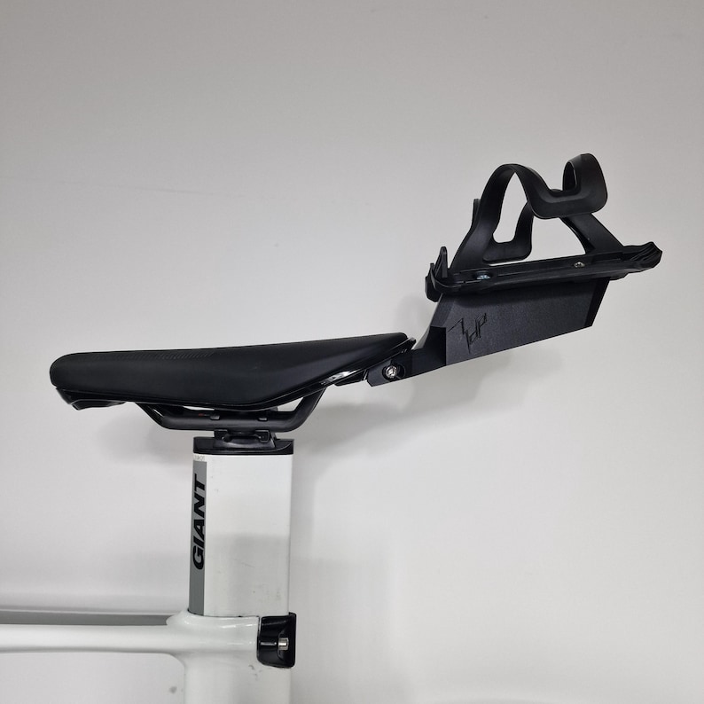 May include: A black bicycle saddle mounted on a white Giant seat post, featuring an attached black rear bottle cage mount with a stylized logo. The assembly includes a matte black water bottle cage secured to the rear bracket behind the seat.