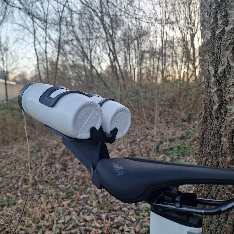 May include: A black bicycle saddle mount featuring two white water bottles held in place by black plastic cages. The mount is attached to the rear of a black bicycle seat, with the bottles positioned horizontally behind the saddle.