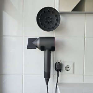 May include: A black hair dryer with a pink accent is mounted on a white tiled wall. A black diffuser attachment is also mounted on the wall. The hair dryer is plugged into an electrical outlet.