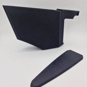 May include: A black plastic 3D printed phone stand with a small, separate black plastic piece. The stand has a raised edge on the back and a small handle on the side. The stand has the word "Zero" printed on the front.