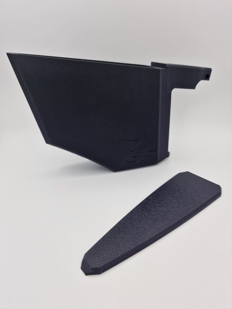 May include: A black plastic 3D printed phone stand with a small, separate black plastic piece. The stand has a raised edge on the back and a small handle on the side. The stand has the word "Zero" printed on the front.