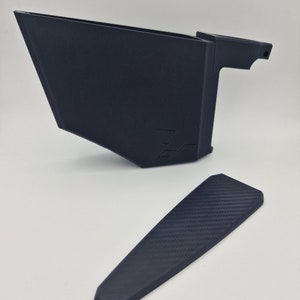 May include: A black plastic 3D printed phone stand with a carbon fiber pattern on the base. The stand has a raised lip on the back to prevent the phone from sliding off.