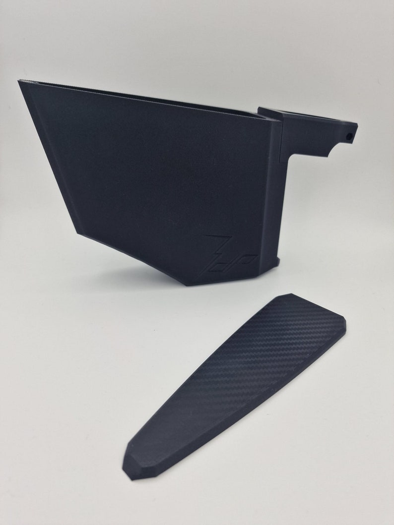 May include: A black plastic 3D printed phone stand with a carbon fiber pattern on the base. The stand has a raised lip on the back to prevent the phone from sliding off.