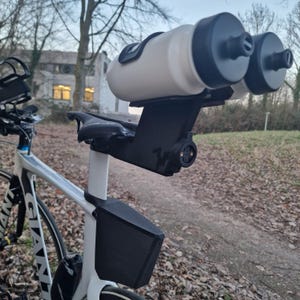 May include: A white bicycle frame featuring a black rear-mounted dual water bottle holder attached to the seat post. The system includes two white water bottles with black caps, a black storage box mounted below the saddle, and a black bike seat.