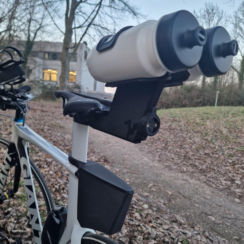 May include: A white bicycle frame featuring a black rear-mounted dual water bottle holder attached to the seat post. The system includes two white water bottles with black caps, a black storage box mounted below the saddle, and a black bike seat.