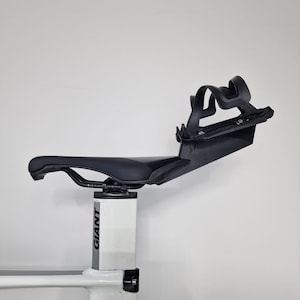 May include: A black bicycle saddle mounted on a white seat post with the word GIANT printed vertically in black. A black plastic water bottle cage is attached to the rear of the saddle, extending upwards and backwards from the seat base.