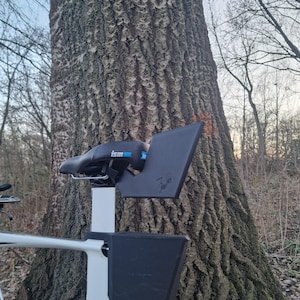 May include: A black ISM bicycle saddle mounted on a white seat post, featuring a black plastic rear storage attachment with the text 'ism' in blue and white, and a signature etched into the side of the black plastic accessory component.