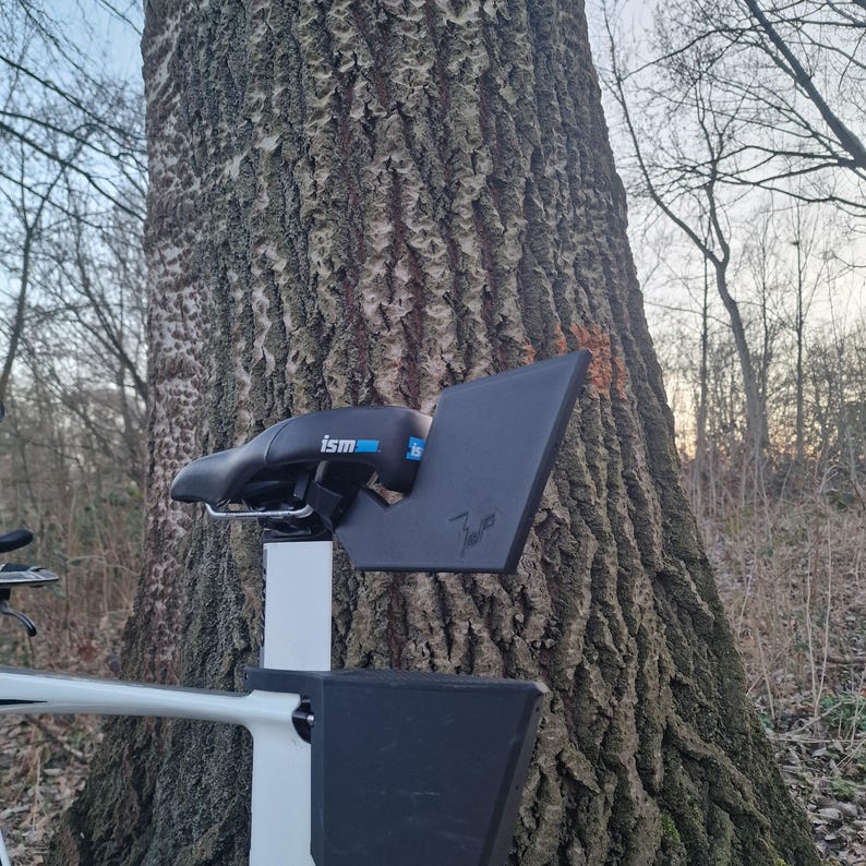 May include: A black ISM bicycle saddle mounted on a white seat post, featuring a black plastic rear storage attachment with the text 'ism' in blue and white, and a signature etched into the side of the black plastic accessory component.
