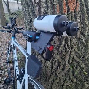 May include: A white and black bicycle featuring a rear-mounted black plastic hydration system holding two white water bottles with black caps. The setup includes a black ISM saddle, a small red rear light, and a black frame storage compartment.