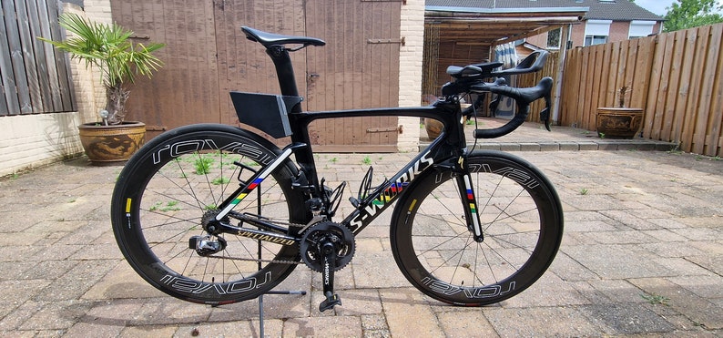 May include: A black Specialized S-Works road bike with a black and white Roval wheelset. The bike has a black frame, black handlebars, and a black seat. The bike is on a stand and has a black water bottle cage.