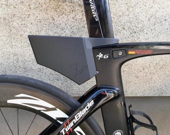 May include: Close-up of a black road bicycle with a black storage box attached to the seat post. The bicycle has a black frame, a black wheel, and the words "TwinBlade" are visible on the frame. The bicycle is against a light gray wall.