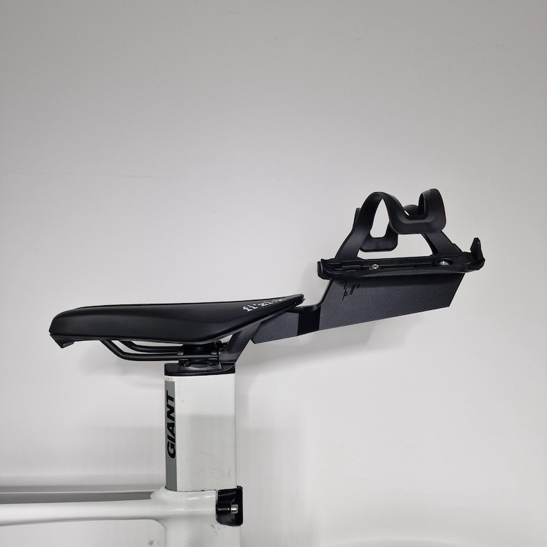 May include: A black bicycle saddle mounted on a white seat post with the brand name GIANT printed in black. Attached to the rear of the saddle is a black metal bracket holding a black plastic water bottle cage, extending horizontally outward.