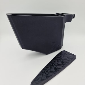 May include: A black 3D printed object with a textured surface and a handle. The object is in two pieces, one is a container and the other is a lid. The lid has a geometric pattern.