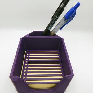 May include: A purple desk organizer with two pens and a notepad. The organizer has a compartment for pens and a tray for the notepad. The notepad has yellow pages with horizontal lines.