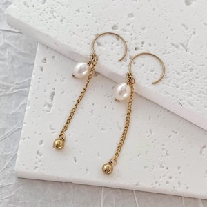 May include: A pair of gold hoop earrings with a delicate chain and a pearl dangling from each hoop.