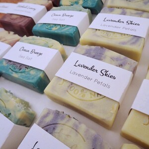 Puede incluir: Surtido de jabones artesanales con varios aromas y colores. Las barras de jabón están etiquetadas con el aroma y los ingredientes, incluyendo "Ocean Breeze", "Lavender Skies" y "Sin perfume".