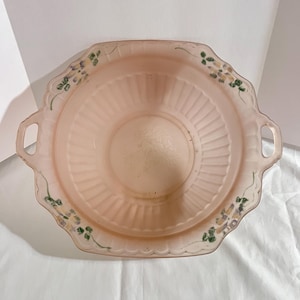 May include: A vintage pink glass serving bowl with a scalloped edge and two handles. The bowl features a ribbed interior and is decorated with painted floral accents in green, yellow, and purple. The bowl is likely from the mid-20th century.