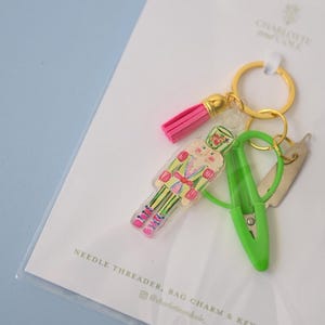 May include: A keychain set on a white display card labeled "NEEDLE THREADER, BAG CHARM & KEYCHAIN" and "CHARLOTTE AND COLE". The set includes a gold ring, a pink tassel, a clear acrylic nutcracker charm, a bright green needle threader, and a metal tool.