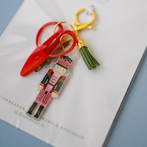 May include: A keychain featuring a clear acrylic nutcracker charm with red, green, and gold details, a pair of small red metal scissors, and a green faux leather tassel. The components are attached to a gold-toned metal lobster clasp.