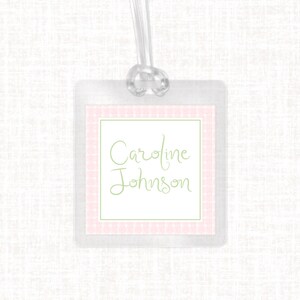 May include: Clear plastic luggage tag with a pink and white gingham border and a green inner border. The name "Caroline Johnson" is written in green script within the inner border. A clear plastic loop is attached at the top.