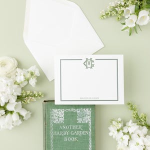 May include: A white envelope with a green and white monogrammed stationery card. The card has a black border and the text "Maeghan Gann". A green book with the title "Another Hardy Garden Book" by Helena Rutherford Ely is in the bottom left corner of the image.